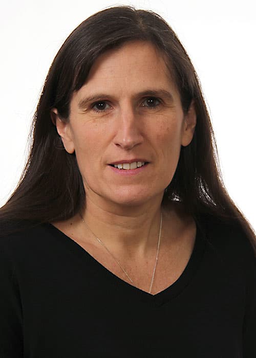 Marie-Noëlle Albert, cheveux longs bruns et haut noir, regarde l'objectif devant un fond clair uni.